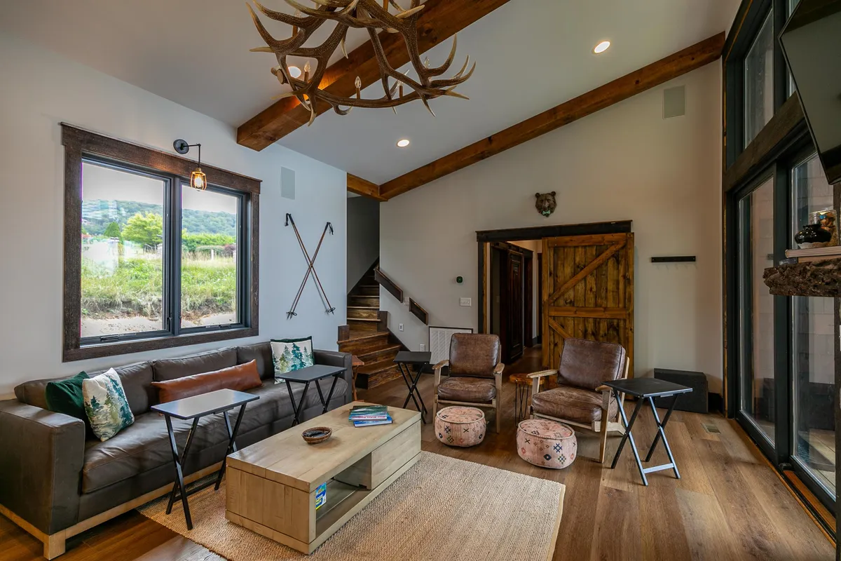 A spacious and airy living area featuring an antler chandelier, a leather sofa, a rustic wooden coffee table, and expansive windows that invite abundant natural light. The large windows offer glimpses of the outdoors, connecting the interior with the surrounding mountain landscape.