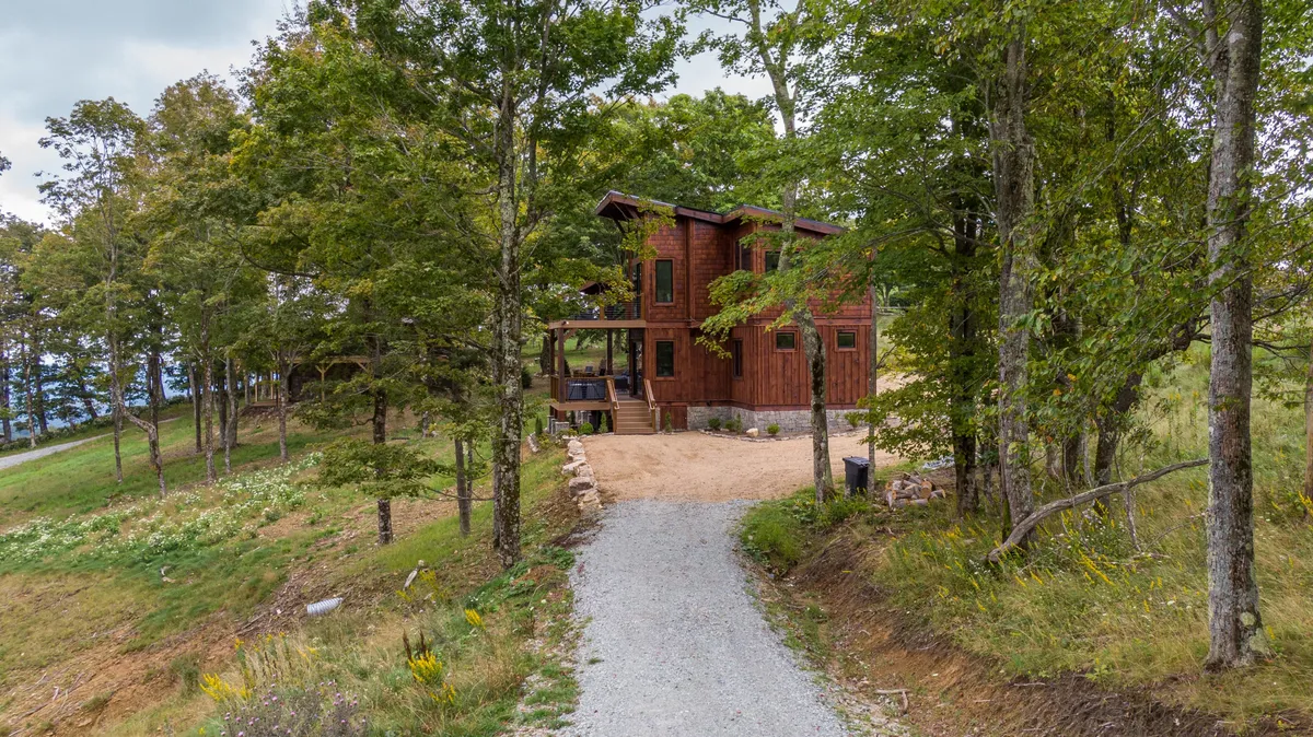 A serene exterior view of the wooden cabin nestled among tall trees, with a gravel pathway leading to the entrance. The surrounding landscape hints at opportunities for outdoor exploration and group activities, reinforcing the sense of adventure and tranquility.