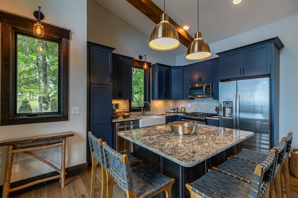 Focusing on the kitchen, this image features a striking granite countertop, modern blue cabinetry, metallic pendant lights, and a large window framing the scenery outside. The kitchen is clearly designed for communal cooking and entertaining, with stylish yet functional touches.