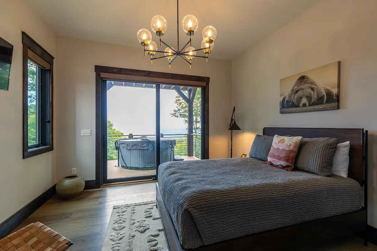 A serene bedroom setting with a bed, elegant chandelier, large windows, and tastefully curated artwork. The windows open to beautiful views, suggesting a seamless transition to a private balcony for witnessing sunrises or stargazing.
