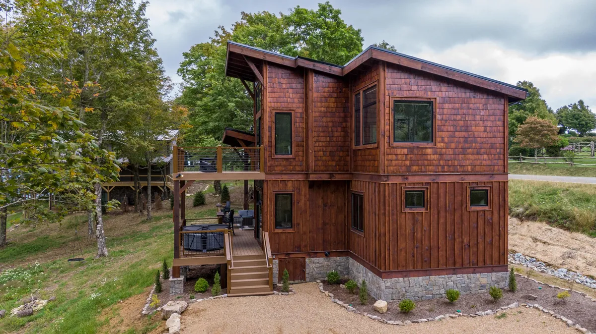 A warm, welcoming exterior shot of the cabin featuring its wooden siding, sturdy stone foundation, inviting deck area, and lush surrounding greenery. The image hints at family-friendly amenities like outdoor dining and evening bonfires, showcasing the cabin as a hub for connection and outdoor fun.