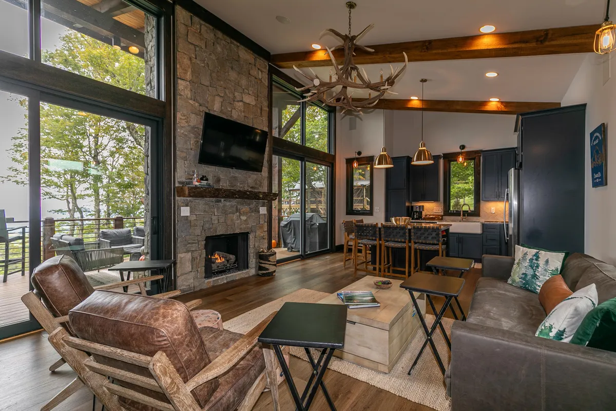 Another angle of the living area, this image highlights the stone fireplace, large windows with breathtaking views, an antler chandelier, exposed wooden beams, and ample, comfortable seating. The room glows with natural light, reinforcing the mountain retreat feel.