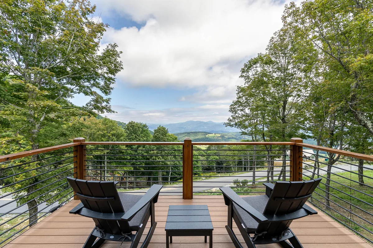 An exterior shot featuring inviting deck chairs arranged on a spacious deck, overlooking a stunning mountain view. The setting is tranquil, with railings framing the panoramic vista and the sky painted with soft light, suggesting either sunrise or sunset. The chairs offer a perfect spot to relax, read, or enjoy the natural landscape.