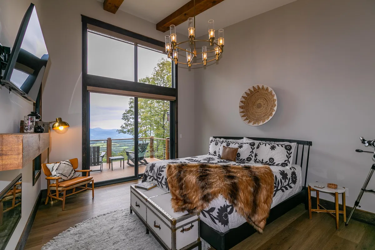 A warmly-decorated bedroom with large windows that frame panoramic views, a stylish chandelier, a comfortable bed dressed in decorative bedding, and wood accents that echo the surrounding landscape. The room feels inviting and closely connected to the natural environment outside.