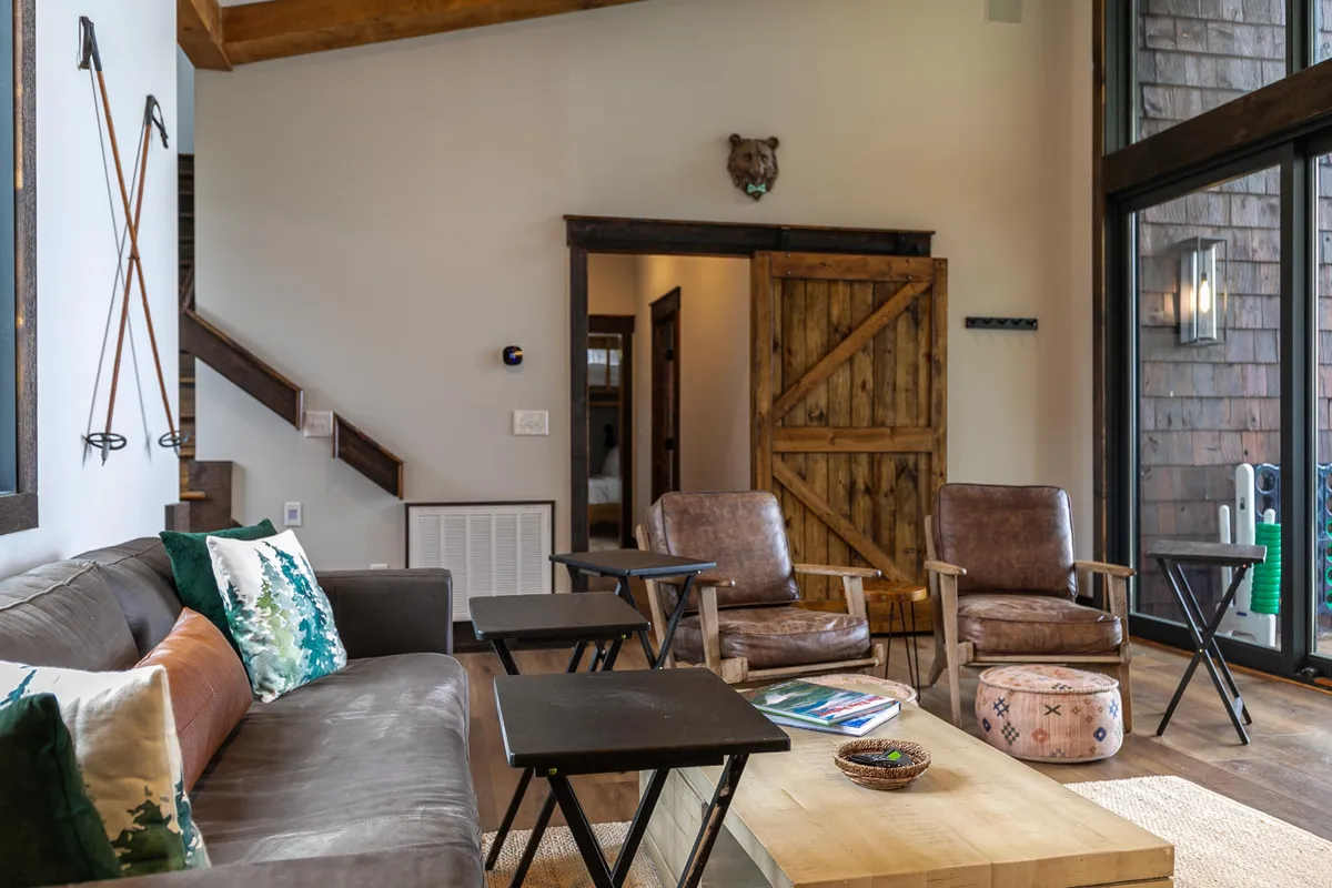 A cozy interior scene with a comfortable sofa adorned with colorful throw pillows, two leather armchairs, and a wooden coffee table with decorative items. Large windows in the background suggest a connection to the outdoors, and the setting is ideal for both relaxation and creative work.