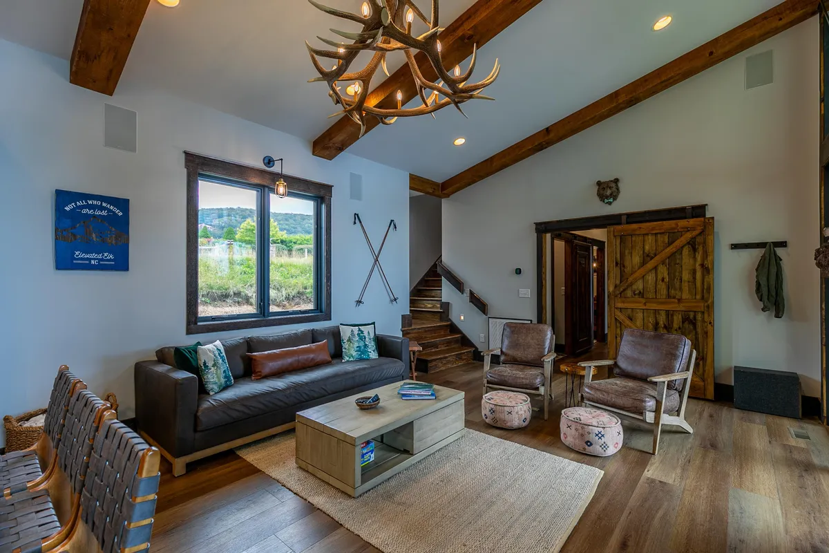 A cozy living space featuring an antler chandelier, large window overlooking the outdoors, a comfortable sofa, and rustic wooden accents. The inviting area encourages relaxation while keeping the stunning mountain setting in full view.