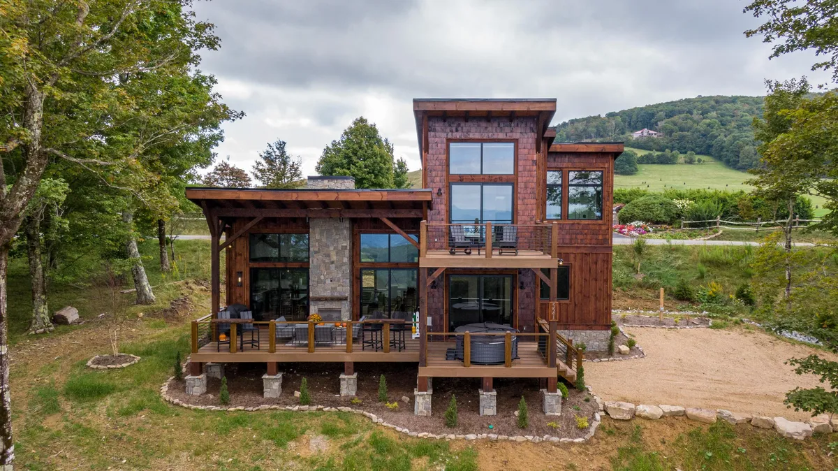 This image showcases the stunning exterior of a cozy mountain cabin nestled among lush greenery. The spacious wooden deck features inviting areas for outdoor relaxation, offering sweeping sunset views. Large windows reflect the natural beauty that surrounds the stone-accented façade, creating a seamless connection between indoors and outdoors.