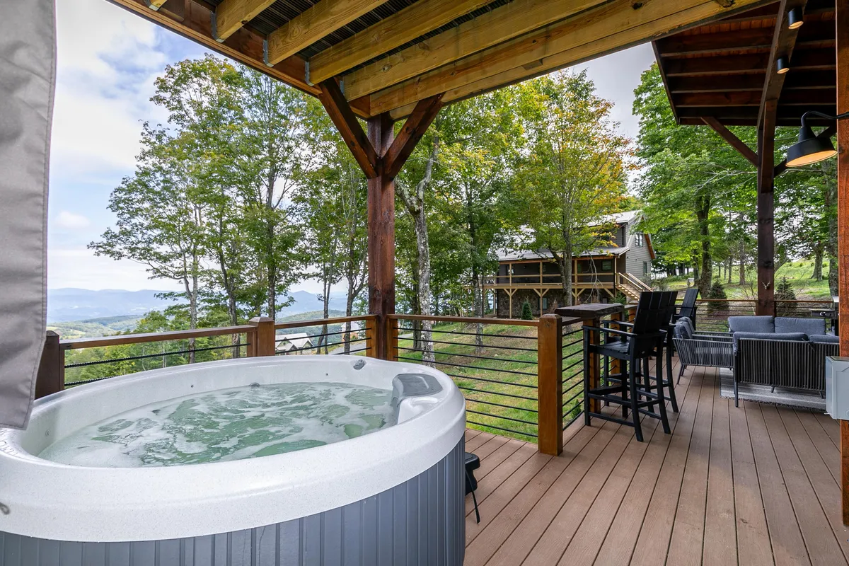 A steaming hot tub is set on a wooden deck, surrounded by tall trees and looking out toward distant mountains. The space is private and infused with tranquil natural beauty, ideal for unwinding after a day of hiking or exploring. The mountains in the distance hint at the surrounding adventure opportunities.