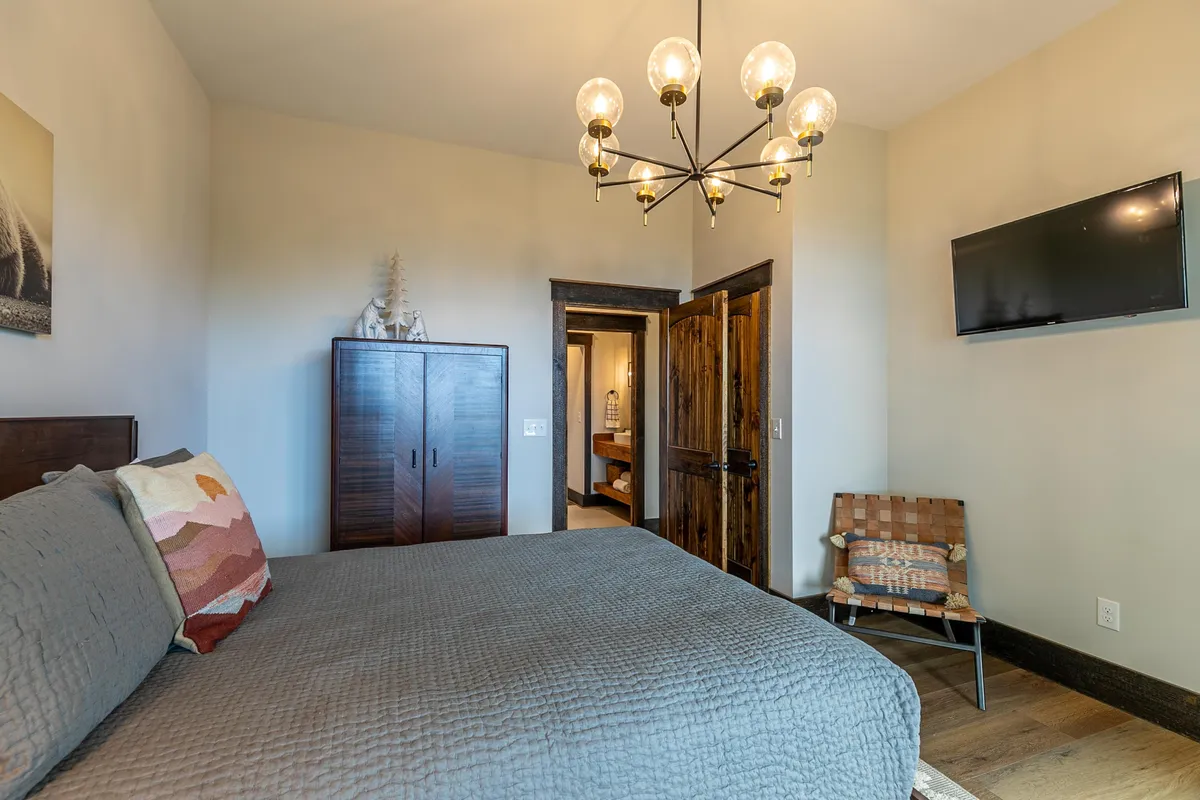 A cozy queen bedroom featuring a comfortable bed with a textured quilt, a stylish chandelier above, a wooden cabinet, and a visible doorway leading to an en-suite bathroom. The room is inviting and designed for relaxation, with soft natural lighting and tasteful decor.