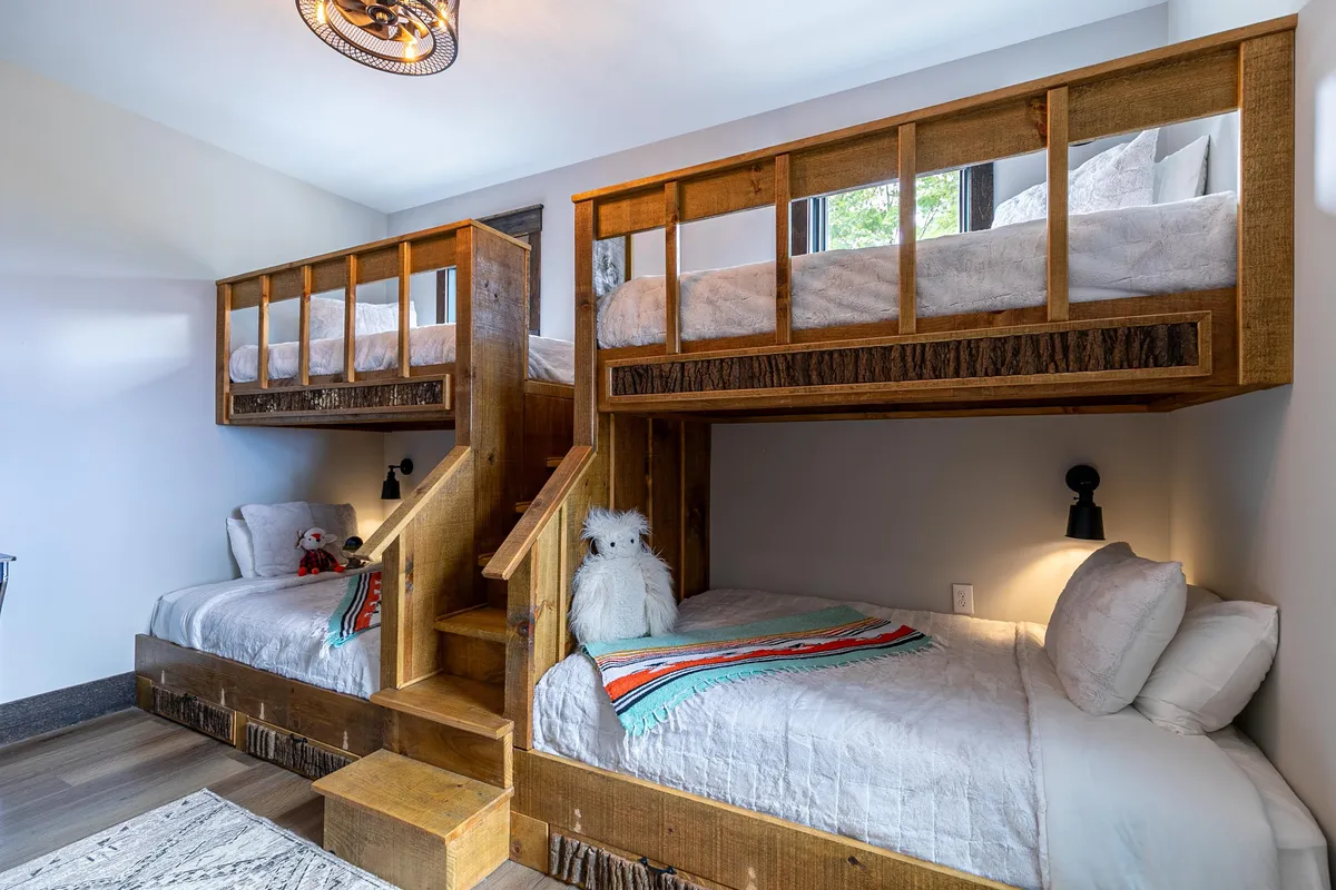 A warm and inviting bunk room equipped with sturdy wooden bunk beds, a staircase for easy access to the top bunks, soft stuffed animals, and a vibrant, colorful blanket. The space exudes a playful, youthful energy perfect for children or groups.