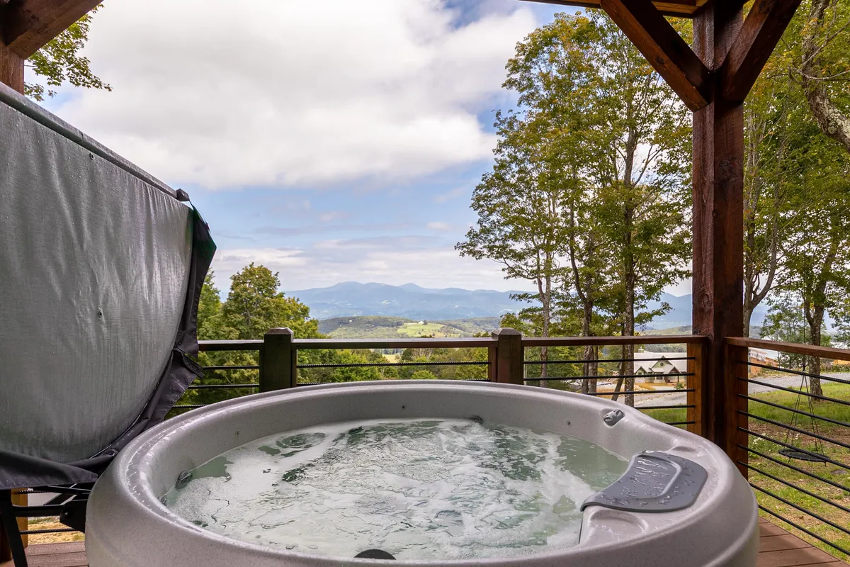 A luxurious hot tub sits on the deck, overlooking a sweeping mountain vista and surrounded by tranquil trees. The serene setting is made for soaking, stargazing, or simply unwinding with nature all around.