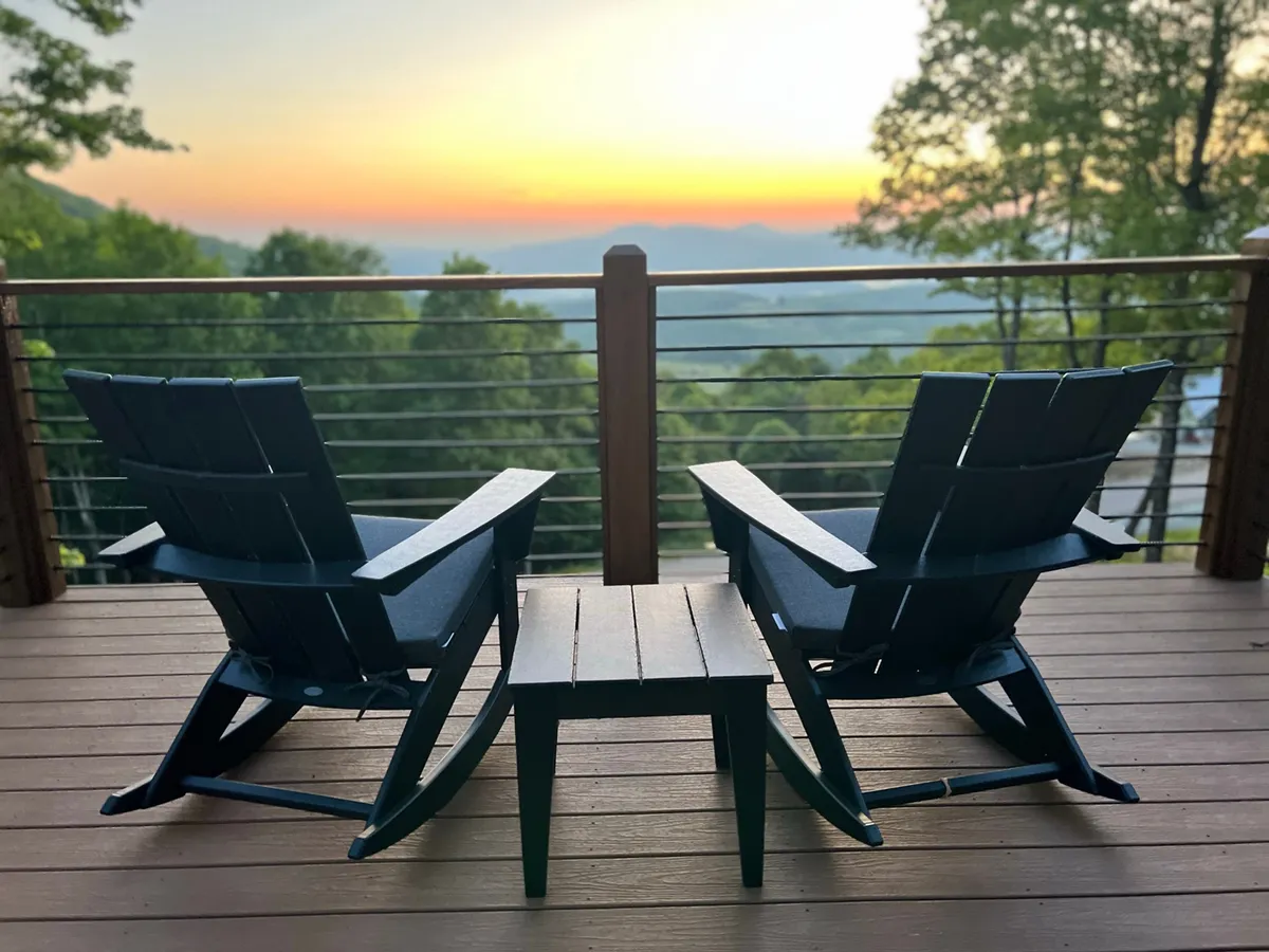 Two Adirondack chairs and a small table are set on a deck overlooking a panoramic mountain vista under a vibrant sunset sky. The setup invites couples to share a quiet evening, savor a drink, or simply enjoy the peaceful mountain air together.
