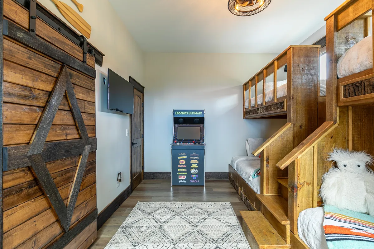 A lively, fun bunk room featuring bunk beds, an arcade machine, a wooden sliding door, and a decorative rug. The presence of the arcade machine injects a sense of excitement and entertainment for kids and teens.