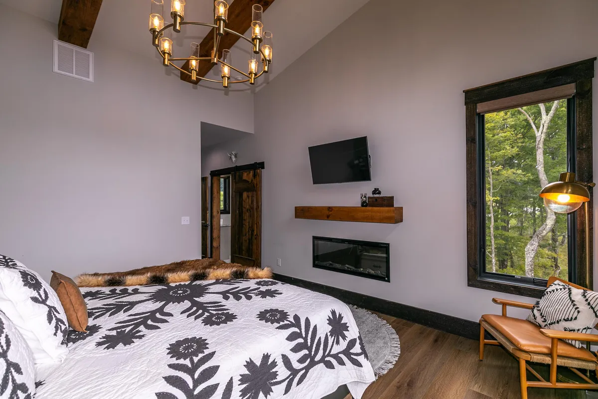 A tranquil master bedroom featuring a bed with a patterned quilt, a cozy fireplace, a large window offering scenic views, and a chandelier overhead. The layout is intimate, inviting relaxation and warmth, while the window frames the mountainous landscape outside.