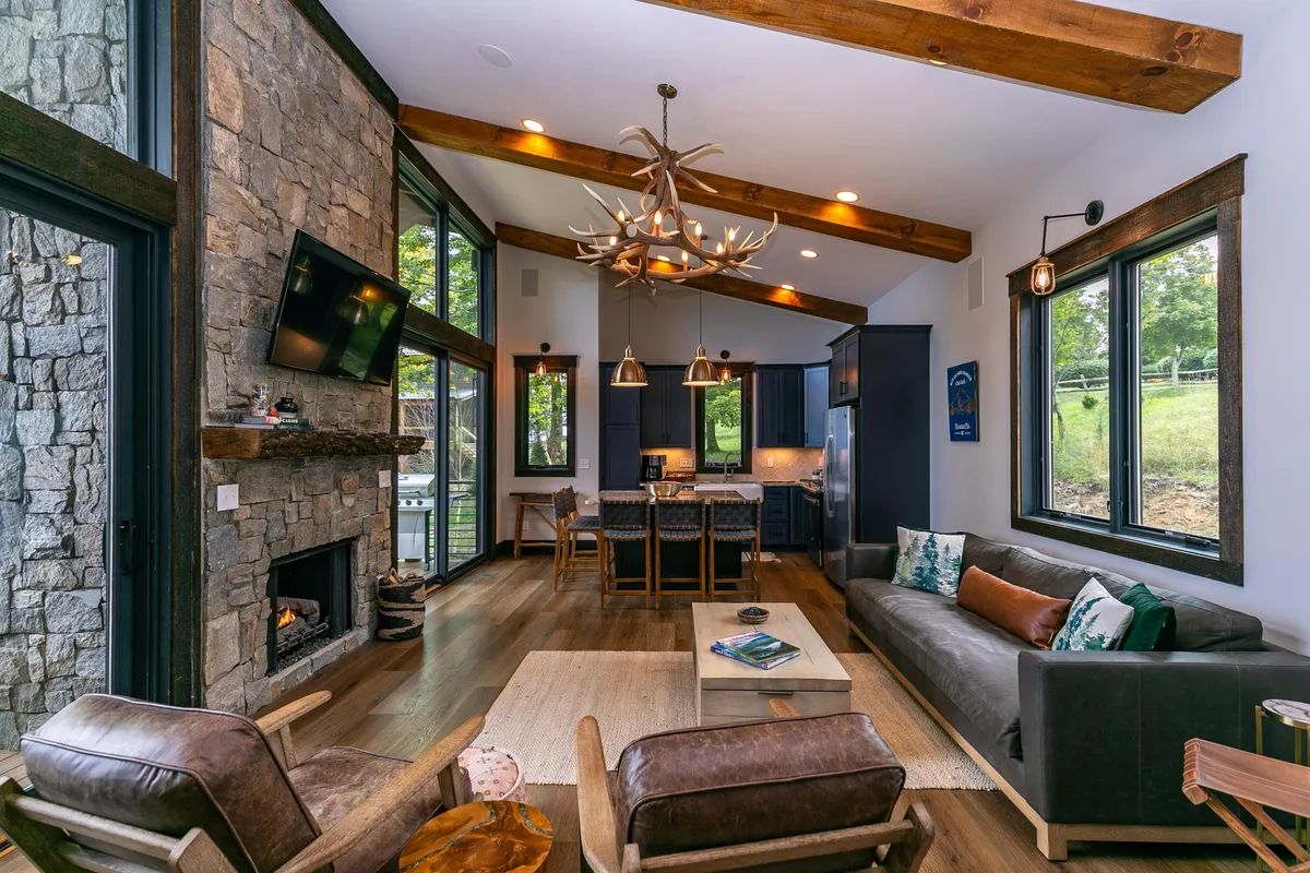 A cozy interior scene focused on a stone fireplace, large windows that flood the space with natural light, an antler chandelier, and plush sofa seating. The room feels inviting, perfect for family gatherings, playing games, or enjoying a movie night after a day of adventure.