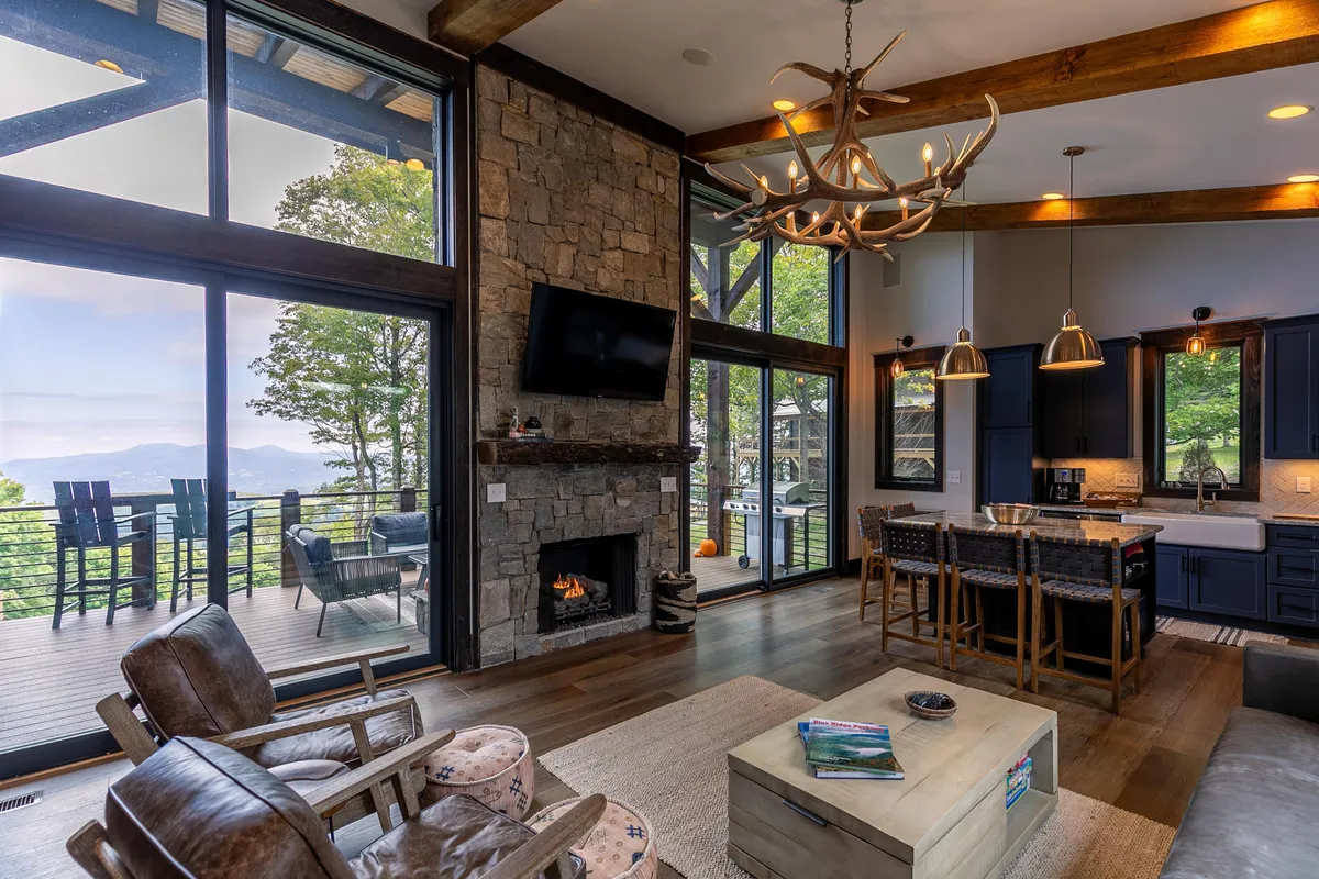 This image showcases the core of the cabin’s open-concept design, with a large stone fireplace as a focal point, comfortable seating arranged for gatherings, an expansive wall of windows offering sweeping mountain views, a kitchen island with seating, and a rustic antler chandelier above. The décor blends natural wood elements with modern comforts, and sunlight pours in, highlighting the inviting atmosphere.