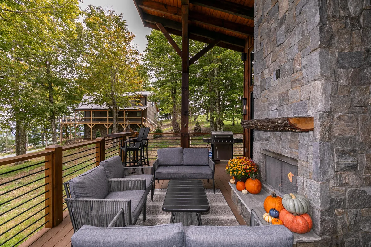 The image captures an outdoor deck with a comfortable seating arrangement in front of a large stone fireplace, a grill nearby, and autumn-themed decorations. The area is styled for both dining and relaxing, set against the backdrop of an expansive deck.